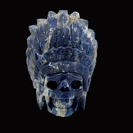 A hand-carved sodalite crystal figurine in the shape of an Indian skull, predominantly blue with natural mineral patterns, displayed on a white stand.